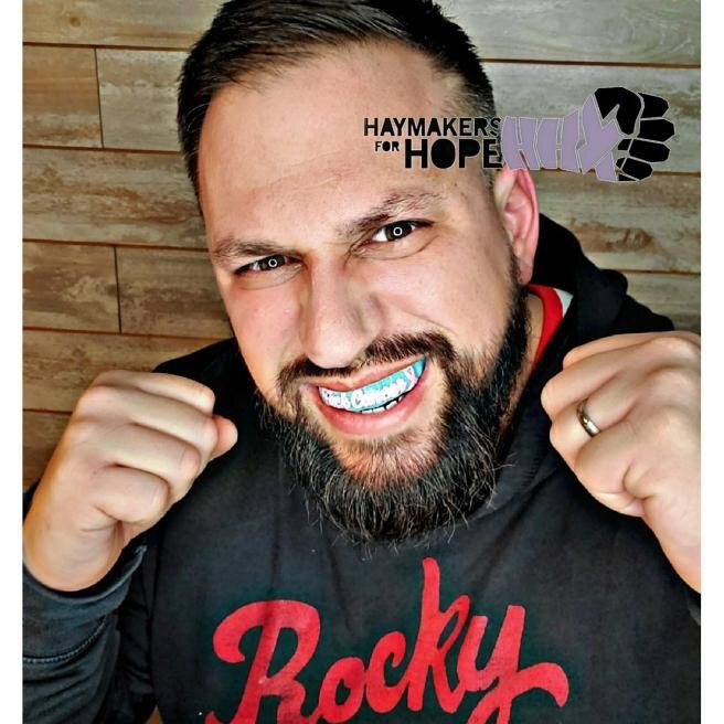 MudHuslters Boxing photo with his hands up, Mouthpiece in front of a wood wall.